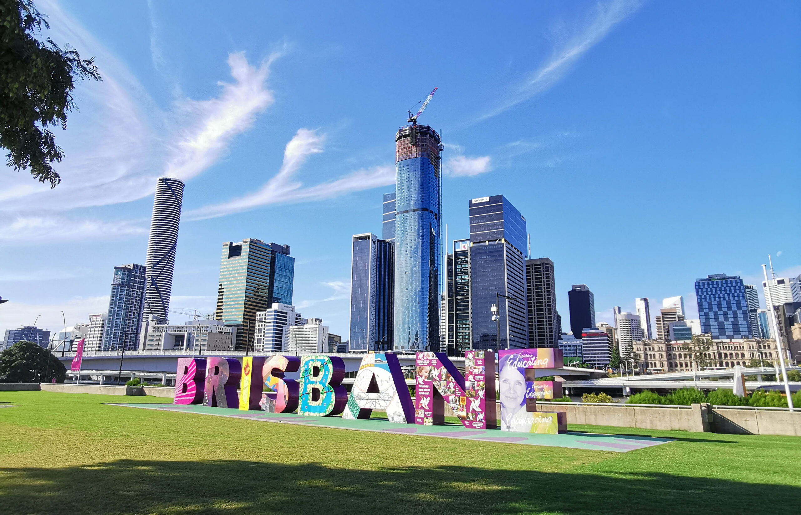 large 3D Brisbane sign sitting on lawn in front of river, with Brisbane city skyline in the background